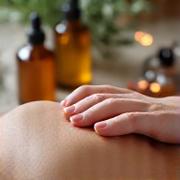 Manos aplicando un aceite esencial en la espalda de una persona, con botellas de aceites e ingredientes naturales borrosos en el fondo, transmitiendo relajación.